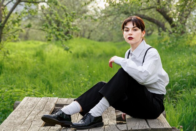 Una hermosa chica con una camisa blanca y pantalones negros con tirantes contra el fondo del cielo y la hierba verde