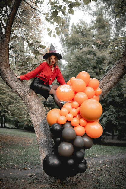 Hermosa chica con camisa naranja, falda negra y sombrero de bruja Se sienta en un árbol Diseño de arte de fiesta de Halloween