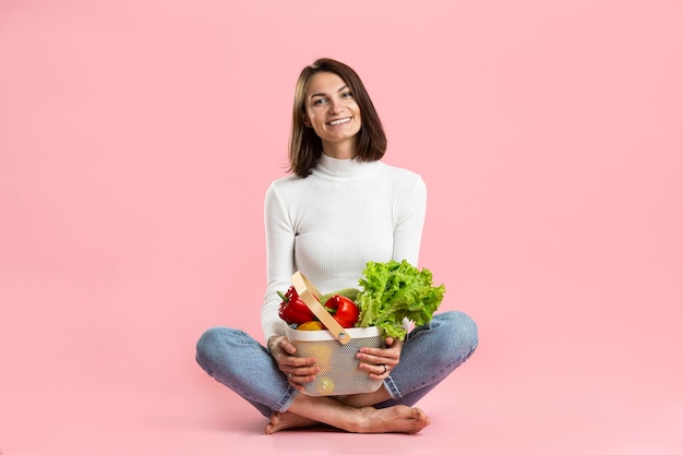 Hermosa chica con una canasta de fibra de vegetales saludables se sienta sobre un fondo rosa