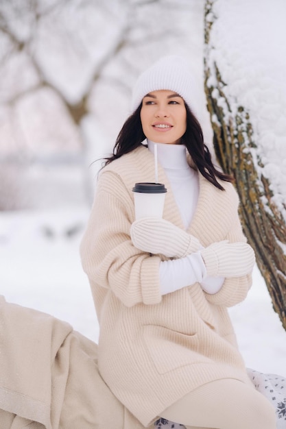 Una hermosa chica con un cárdigan beige y un sombrero blanco disfrutando de beber té en un bosque de invierno nevado cerca de un lago