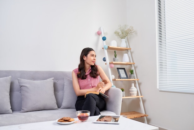 Hermosa chica en casa sentada en el sofá, leyendo un libro y tomando un café