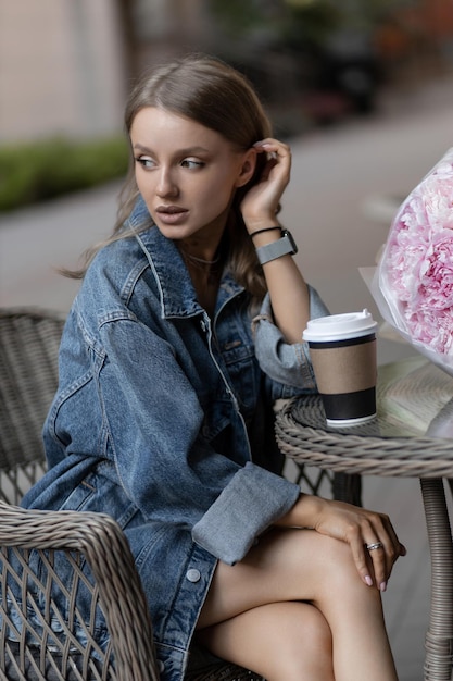 Una hermosa chica caucásica con un atuendo informal se sienta en un café de la calle y bebe café junto a hermosas flores de peonía