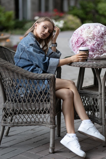 Una hermosa chica caucásica con un atuendo informal se sienta en un café de la calle junto a hermosas flores de peonía