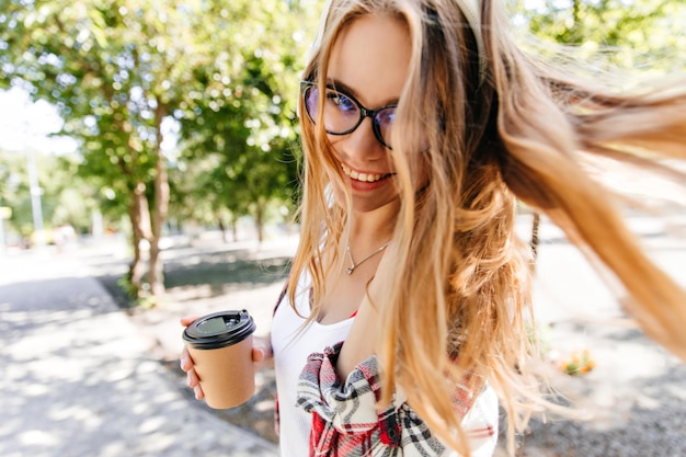 Hermosa chica caucásica con cabello largo bebiendo café con leche en el parque Maravillosa dama sonriente en vasos disfrutando de café en el fondo de la naturaleza