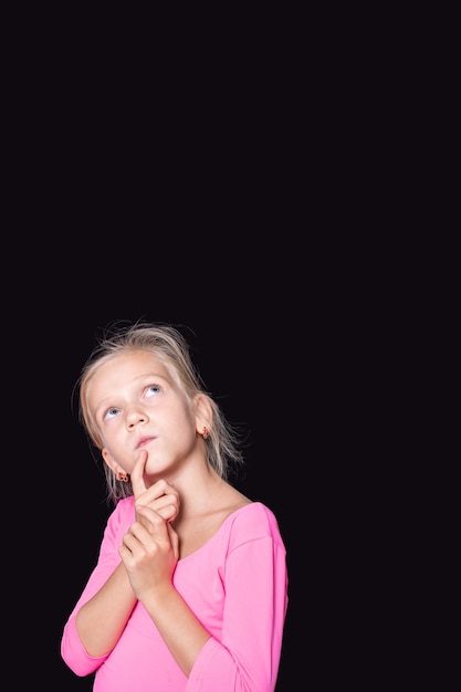 Hermosa chica caucásica pensativa en un suéter rosa mirando hacia arriba y sosteniendo su dedo a la cara sobre un fondo negro, copie el espacio. Una plantilla para superponer su texto.