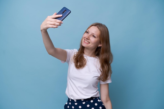 Hermosa chica caucásica tomando una foto selfie fotografiándose en un teléfono inteligente hablando en una videollamada en línea con una camiseta blanca con el pelo largo aislado en un fondo azul