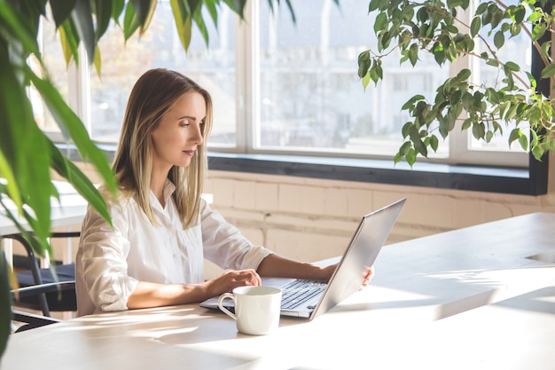 Hermosa chica caucásica trabajando en una computadora portátil de forma remota en un espacio luminoso con plantas verdes.