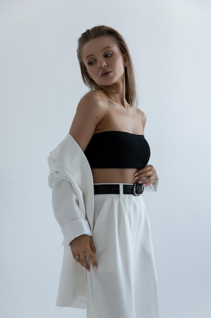 Una hermosa chica caucásica con un traje blanco se para en una pose de moda en un estudio con un fondo blanco y se lo quita