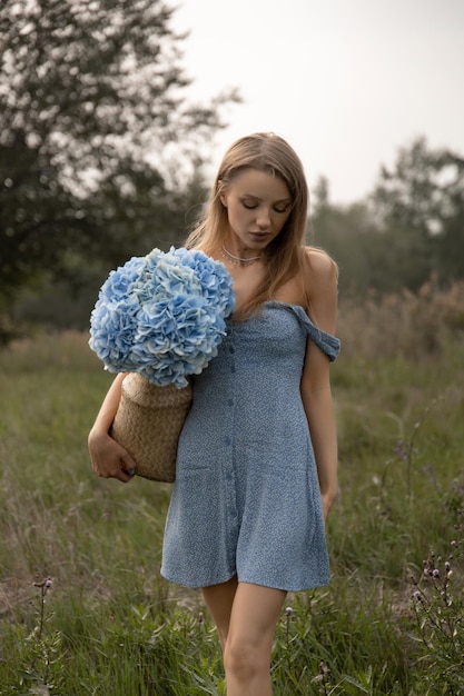 Hermosa chica caucásica con un vestido azul camina por el campo con una canasta de flores