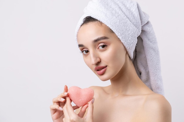 Hermosa chica con cejas gruesas y piel perfecta en toalla de fondo blanco en la cabeza foto de belleza Sosteniendo una esponja de corazón rosa cosmética