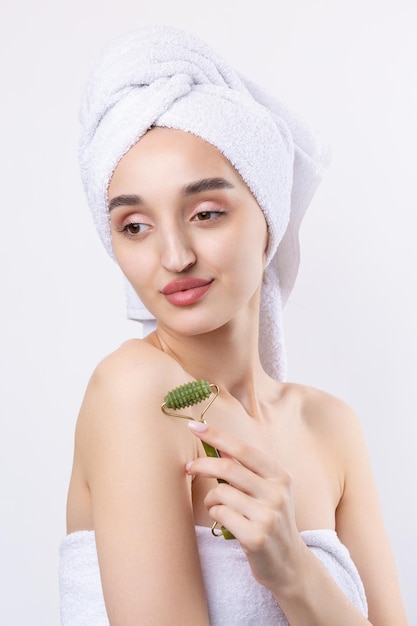 Hermosa chica con cejas gruesas y piel perfecta en una toalla de fondo blanco en la foto de belleza de la cabeza Sostiene un rodillo cosmético