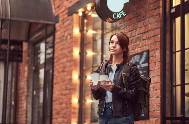 Una hermosa chica con una chaqueta de cuero con una mochila sosteniendo tazas con café para llevar afuera cerca del café.