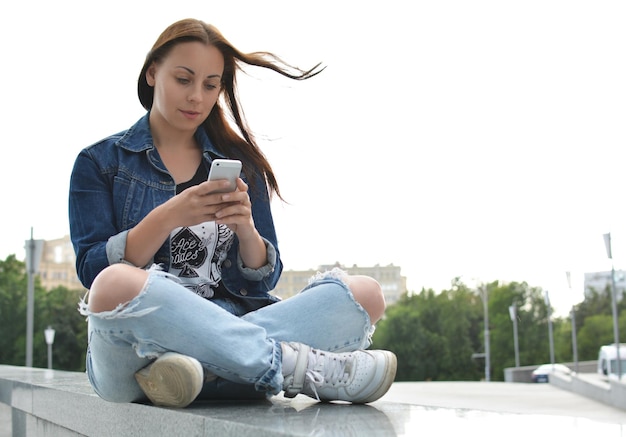 Una hermosa chica con una chaqueta de jeans sentada en los escalones de una calle de la ciudad usa su teléfono inteligente