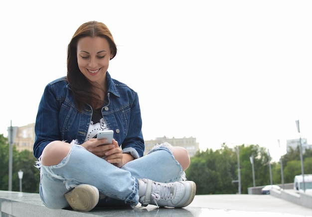 Una hermosa chica con una chaqueta de jeans sentada en los escalones de una calle de la ciudad usa su teléfono inteligente