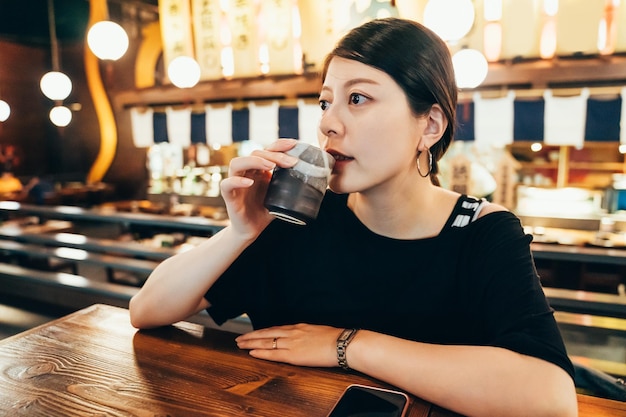 Hermosa chica china asiática bebiendo té en izakaya. Mujer de belleza se casa con anillos en las manos de los dedos poniendo sobre una mesa de madera por teléfono celular. dama con una taza de bebida caliente en un restaurante japonés