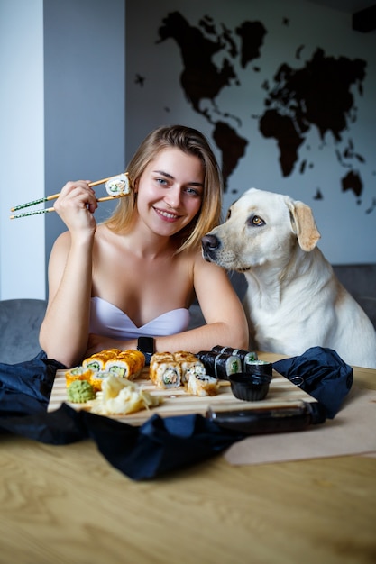 Hermosa chica comiendo primer plano de sushi con su perro labrador, sonriendo, sosteniendo un rollo de sushi con palillos. Comida japonesa saludable.