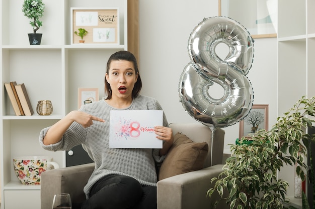 Hermosa chica confundida en el día de la mujer feliz sosteniendo y puntos en la tarjeta de felicitación sentado en un sillón en la sala de estar