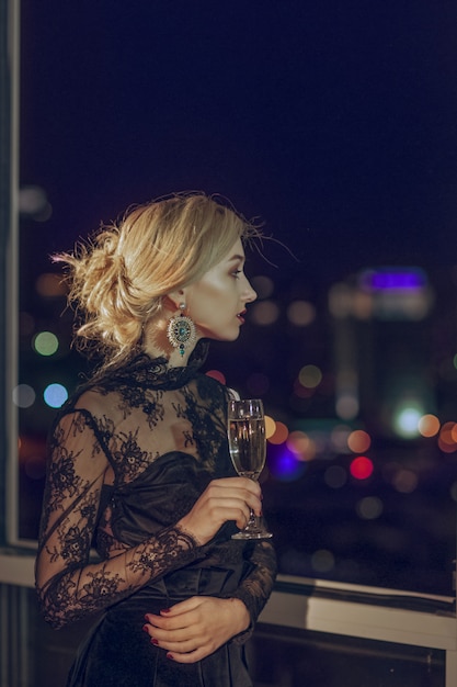 Hermosa chica con copa de champán de pie cerca de la ventana con la ciudad de la noche borrosa