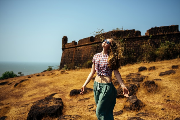Hermosa chica delgada en una camisa y gafas de sol en una colina contra el mar y el antiguo fuerte portugués en Goa. Mujer joven con cabello suelto gira su rostro hacia el sol y disfruta de la vida.