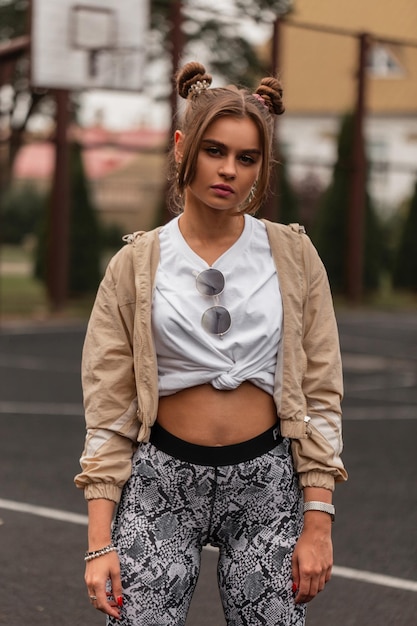 Hermosa chica deportiva de moda en ropa deportiva elegante con sudaderas con capucha y pantalones de chándal en el patio de recreo