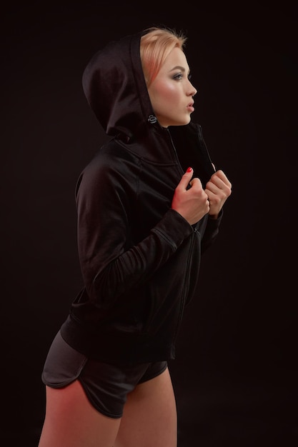 Hermosa chica deportiva seductora con la figura delgada y sexy en la sudadera con capucha negra, ropa interior deportiva y pantalones cortos negros está posando en el estudio