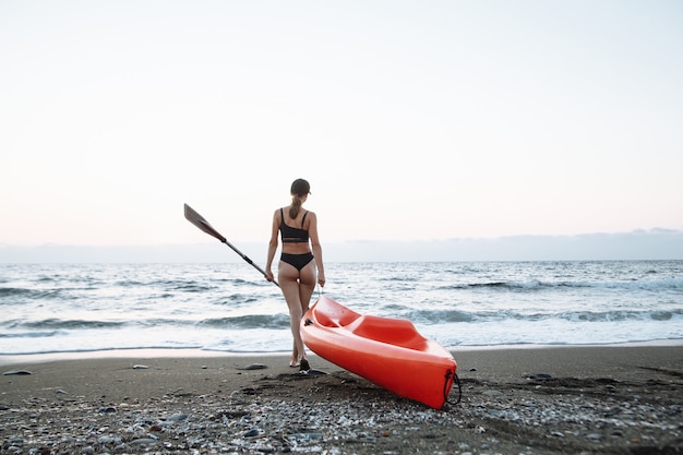 Hermosa chica deportiva en traje de baño negro va con un kayak naranja para nadar en el mar al atardecer