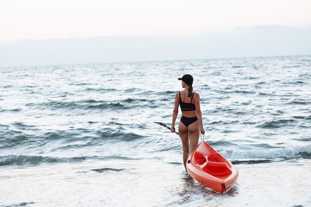 Hermosa chica deportiva en traje de baño negro va con un kayak naranja para nadar en el mar al atardecer