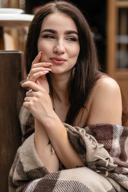 Hermosa chica descansando sentado en la cafetería en la terraza, envuelto en una manta de cuadros de lana. Mañana de otoño. Hermosa mujer joven con una taza de café al aire libre en el día de otoño