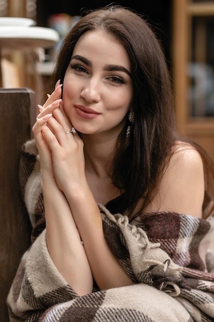 Hermosa chica descansando sentado en la cafetería en la terraza, envuelto en una manta de cuadros de lana. Mañana de otoño. Hermosa mujer joven con una taza de café al aire libre en el día de otoño