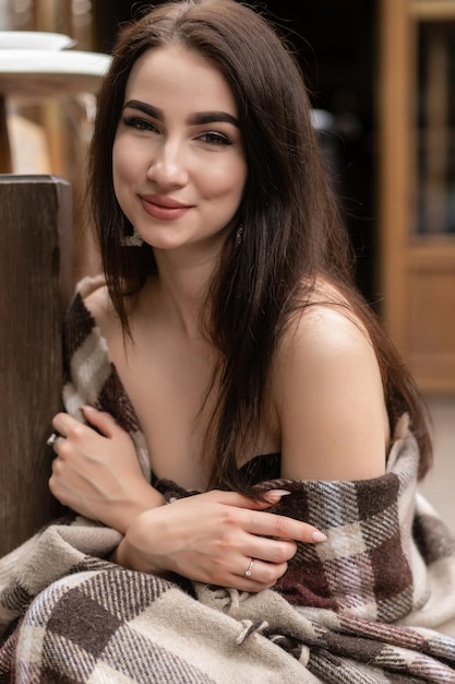 Hermosa chica descansando sentado en la cafetería en la terraza, envuelto en una manta de cuadros de lana. Mañana de otoño. Hermosa mujer joven con una taza de café al aire libre en el día de otoño