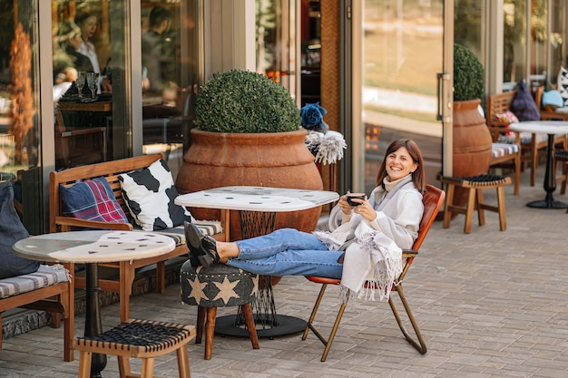 Hermosa chica descansando y tomando café sentado en la cafetería en la terraza, envuelto en una manta de cuadros de lana. Mañana de otoño.