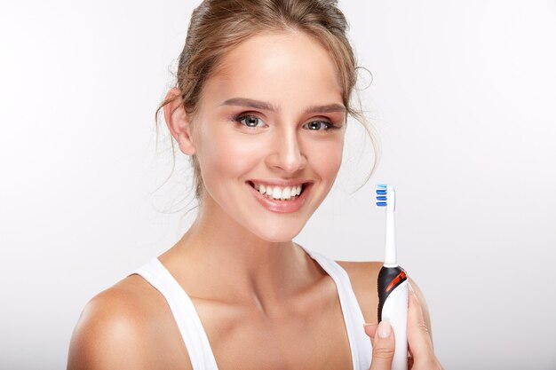 Hermosa chica con dientes blancos como la nieve sobre fondo blanco de estudio, concepto de odontología, sonrisa perfecta, sosteniendo cepillo de dientes eléctrico, espacio de copia.