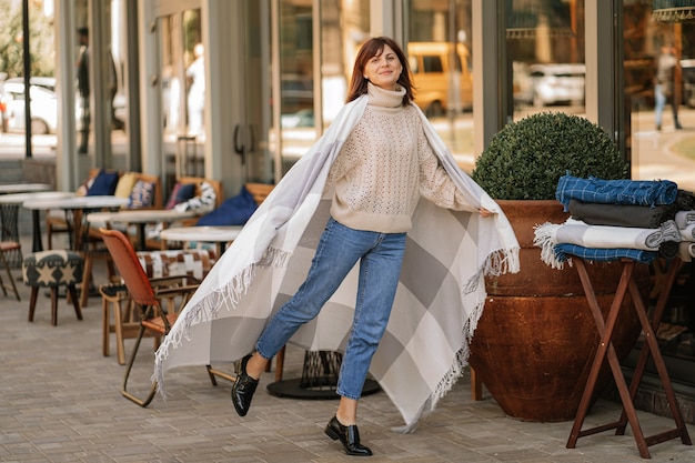 Hermosa chica divirtiéndose caminando en la cafetería en la terraza, envuelta en una manta de lana a cuadros. Mañana de otoño.