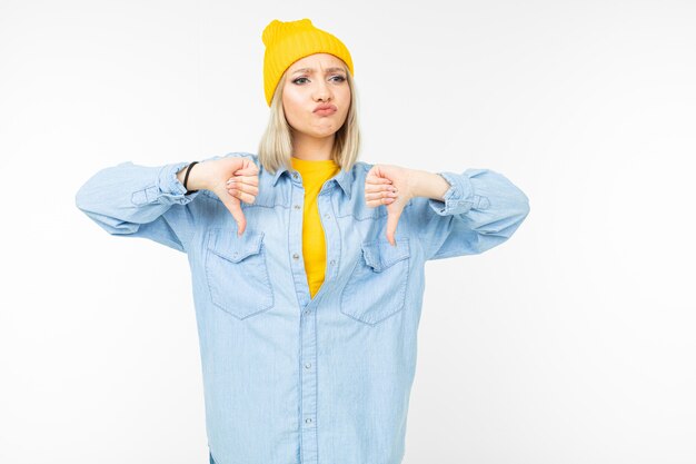 Hermosa chica en una elegante camisa de mezclilla y un sombrero amarillo muestra disgusto en un blanco