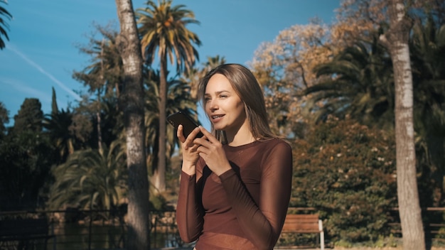 Hermosa chica elegante grabando mensajes de voz en el teléfono inteligente en el parque