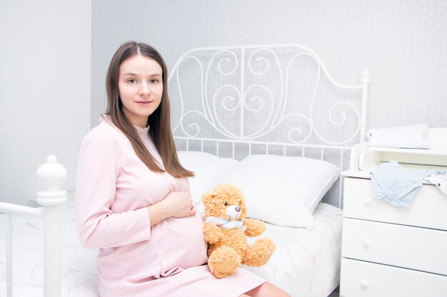 Hermosa chica embarazada está sentada en la cama y sus manos están sobre su estómago. concepto de embarazo, maternidad, preparación y expectativa.