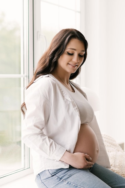 Hermosa chica embarazada con rizos en una camisa blanca y jeans en un estudio luminoso con un interior elegante el concepto de embarazo feliz y familia