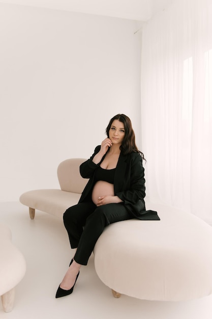 Hermosa chica embarazada con rizos en un traje negro sobre un fondo blanco el concepto de un feliz embarazo y familia