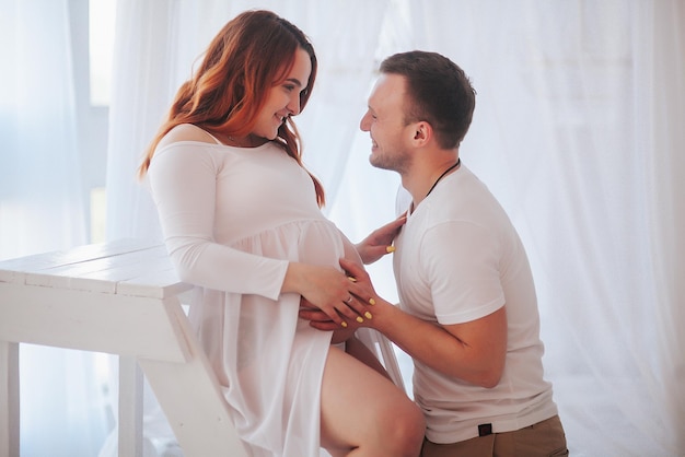 Hermosa chica embarazada en vestido blanco con marido en estudio