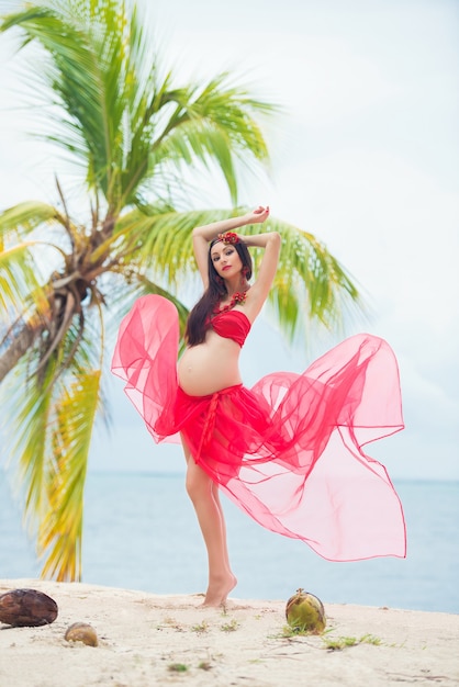 Hermosa chica embarazada en vestido rojo en la playa de arena con palmeras