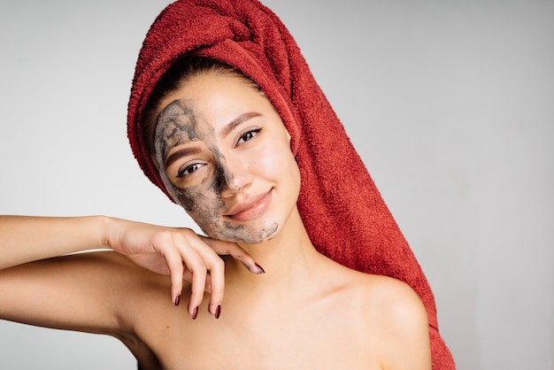 Hermosa chica encantadora con una toalla roja en la cabeza aplicó una útil mascarilla de arcilla a la mitad de la cara, spa de día