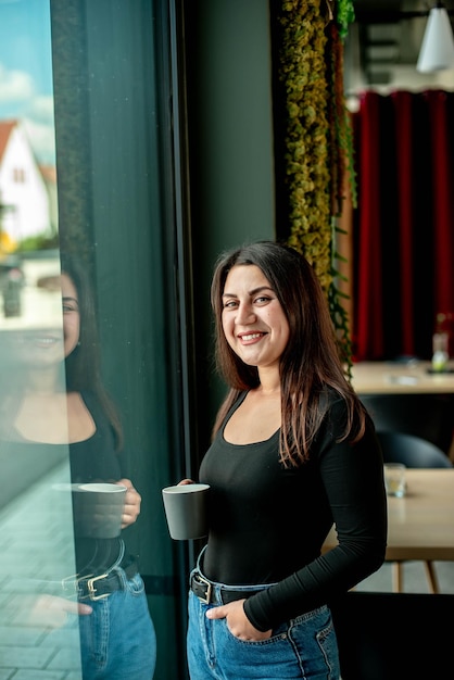 Hermosa chica se encuentra cerca de la ventana con café y sonrisas