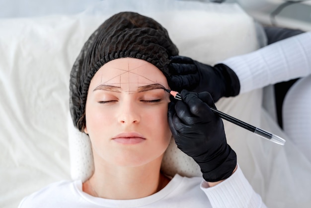 Hermosa chica se encuentra mientras el maestro de maquillaje permanente dibuja la forma de las cejas con lápiz