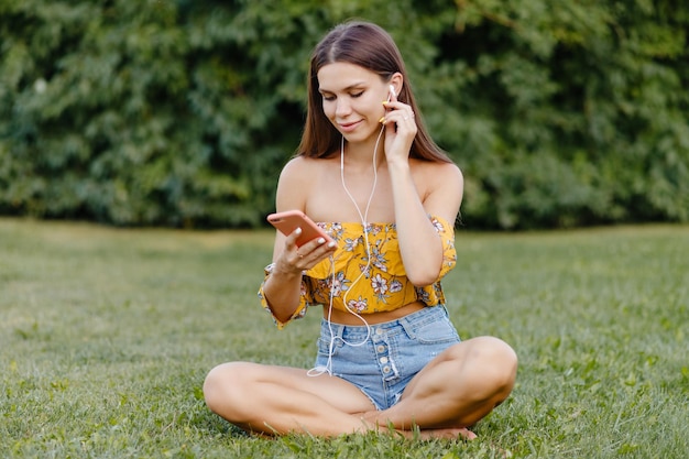 Hermosa chica escucha música en el teléfono mientras se sienta en el césped en el parque