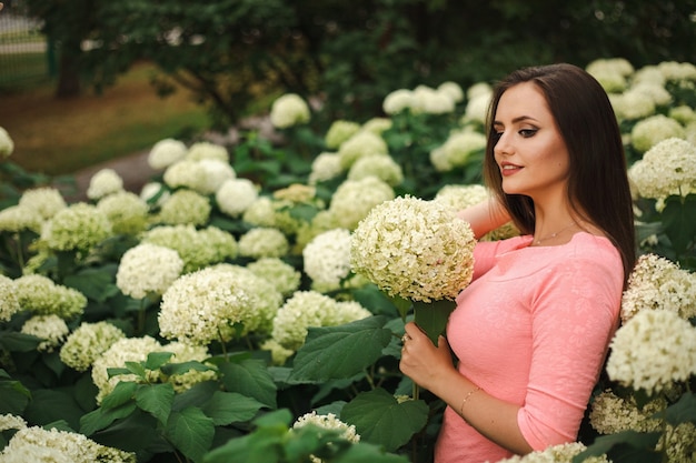 Hermosa chica está en arbustos de flores de hortensias en el jardín al atardecer. Flores en las calles de la ciudad.