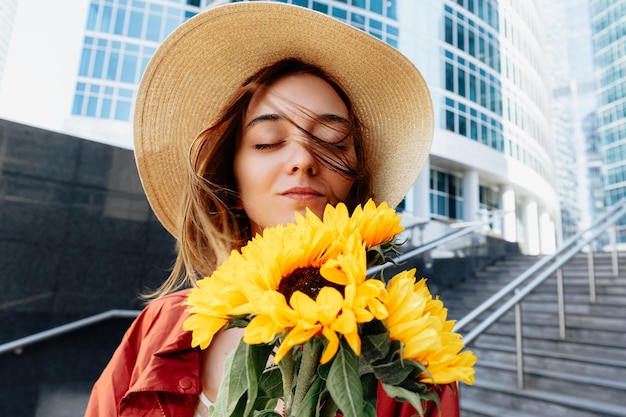 Hermosa chica está oliendo un ramo de flores de girasoles Cabello ondeando en el viento Momento sensual Retrato de mujer de moda urbana al aire libre Concepto de naturaleza en ciudad urbana