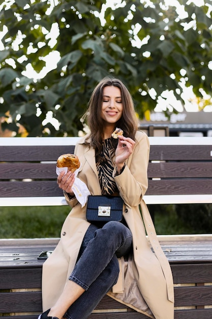 Hermosa chica está sentada en un banco en la calle comiendo un delicioso croissant fresco Una mujer joven rompe un trozo de croissant