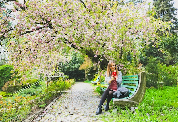 Hermosa chica está sentada en el banco en el jardín bajo el árbol floreciente Blossom sakura al aire libre Estado de ánimo de primavera Mujer en vestido rosa Relajante