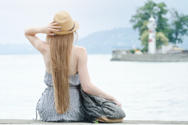 Hermosa chica está sentada en el muelle. El mar y el pequeño faro a lo lejos. Vista trasera