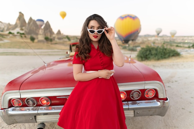 Hermosa chica en estilo retro posando cerca de un auto descapotable rojo vintage en el fondo de globos en Capadocia